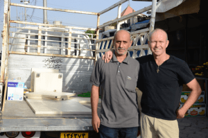 [Image description: Ashur and Father Chris stand next to the bed of the truck, looking at the camera and posing for the photo. They have their arm around the other's shoulders.]